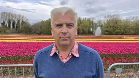 Shaun Whitmore/BBC Mark Eves has white short hair and is wearing  red gingham shirt and a blue V-neck pullover. He is standing in front of rows of different coloured tulips.