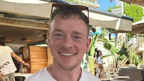 BBC Calum sits in a sunny, outdoor restaurant. He has sunglasses on his head and is smiling. He has blue eyes and two small silver hoop earrings in his left ear. 