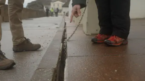 Two people are standing on either side of the long crack in the ground. A person wearing brown hiking boots is stood on the left side of the crack, while a person wearing red walking boots is standing on the right side of the crack. A metal chain leads into the crack. A hand is pointing at the crack.