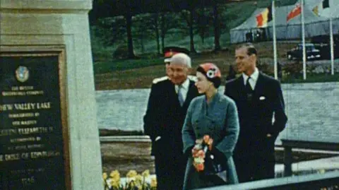 Bristol Water A colour, digitalised image of the late Queen Elizabeth II in a blue, turquoise coat. She is looking at the plaque she's just unveiled saying 'Chew Valley Lake'.  She is stood with her husband, Prince Philip, who is stood behind her in a dark, navy suit and tie. There is a man in a suit talking to them and showing the late Queen and Prince Philip the surroundings. There are flags in the background.