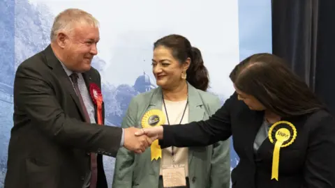 Davy Russel wearing a suit and red rosette shakes the hand of an SNP candidate in a yellow rosette