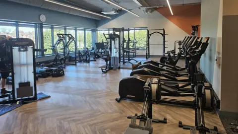 Shropshire Council A gym facility with wood flooring. There are dozens of black fitness machines including rowing machines, ellipticals, and weight lifting equipment. Around the room are large windows which look out onto green trees and housing