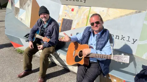BBC/RICHARD EDWARDS This picture shows two friends -  Paul and Dary - busking at Castleford's Tickle Cock Bridge. Dary is on the right of the image, wearing sunglasses, a bodywarmer and a blue shirt. Paul, on the left, has green trousers, a blue jacket and a black beanie hat. Both are smiling and holding acoustic guitars.