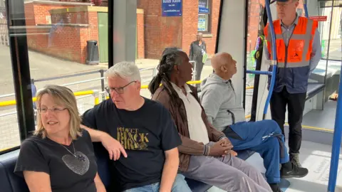 Men and women sit on a Very Light Rail vehicle looking out of the window. One man in a high-vis vest is standing up. There are seats and blue poles for passengers to hold on to.