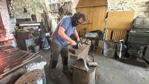 BBC/ Alex James A man in grey trousers and a blue shirt is leaning over an anvil, hammering some metal. He has long brown hair and a big beard. He is wearing safety goggles. To the left of the image is the corner of the smithing fire. 