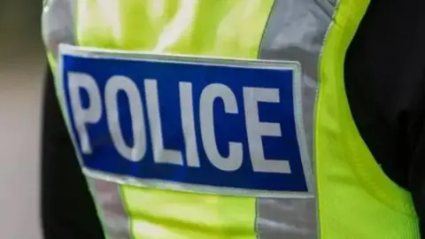 A close-up of a person's back wearing a yellow high-visibility vest with the words "POLICE" written in white and blue across the middle over a black jacket.