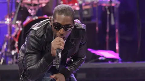 Getty Images Tinie Tempah wears sunglasses and sings into a microphone crouched down. He's wearing a black leather jacket and jeans. He has a short trim hair cut. The background is a band.
