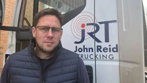 The photo shows Oliver Reid looking at the camera. He is wearing a navy gilet and jumper. He has short brown hair and is wearing glasses. He is standing in front of a white HGV lorry, which has the logo 'John Reid Trucking' on it. 
