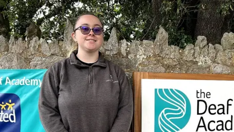 Deaf Academy Hafwen Clarke in a grey jumper and sunglasses looking at the camera