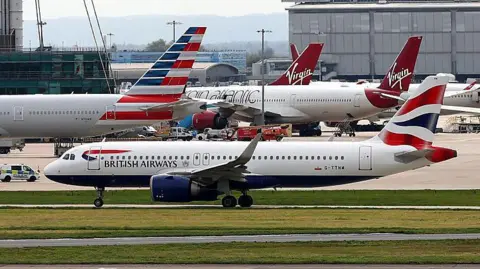 A British Airways plane on the runway with two Virgin Atlantic planes and some other planes behind it. In the background, you can see a Heathrow Airport terminal behind it.