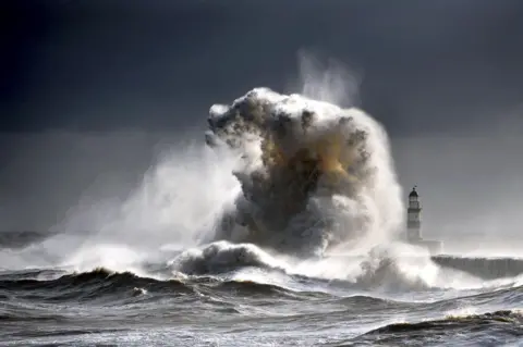 Owen Humphreys/PA Media A giant wave crashes into the pier, towering over the lighthouse. At it's height, the wave is more than double the height of the lighthouse on the left. In the foreground, the waves of the sea are ferocious, whipping up mist and foam. The sky and the sea are so grey the image almost looks like it is in black and white.