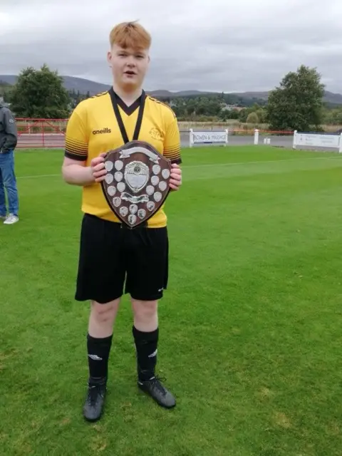 Liz Knox Lewis Knox who played for Fort William Shinty Club in his yellow and black strip holding a wooden trophy covered in small silver shields. 