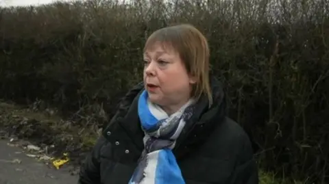 Natalie Farkas, a lady with light brown hair, is wearing a black coat and a blue patterned scarf. She is stood by a hedgerow, with bits of debris from a pile of flytipped pile of rubbish visible.