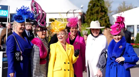 PA Media A line of women laugh as they pose for the camera at the start of Ladies' Day at the 2026 Cheltenham Festival. Some of them have bright yellow or blue outfits on, and all of them are wearing bright fascinators on their heads