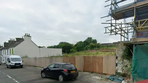 A gap site in Glenluce main street with boards in front of it and a car and a van parked in front of it