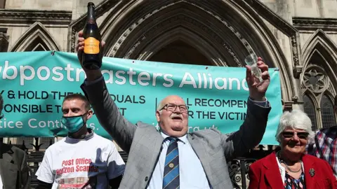 Tom Hedges outside the Court of Appeal after his conviction was quashed. He is wearing a grey suit, sky blue shirt and black and blue striped tie. He has glasses and a white moustache. He holds aloft a bottle of champagne in one hand a glass in the other and is surrounded by other victims in front of a banner.