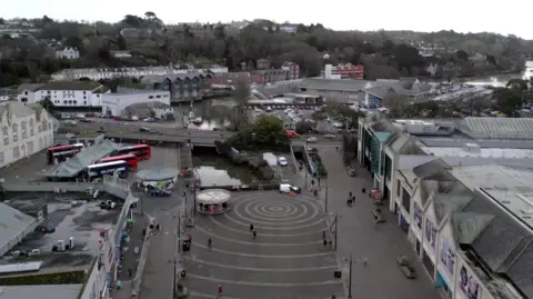 Aerial view of a Truro city centre with circular patterns on the ground, surrounded by buildings, buses and a river with bridges in the background. White skies.