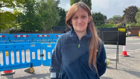 A woman with long dark hair wearing a dark blue fleece. She is stood on a roadside, the road is closed off with blue fencing