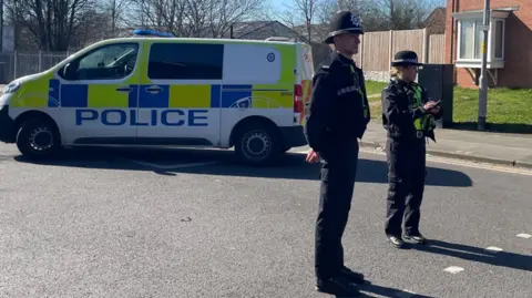 BBC Two police officers stand in the road with a van behind them and police tape in place to their right.