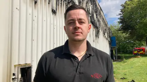 Alex Pope/BBC A man with short brown hair and a black polo shirt is stood in front of a burnt building with a fire engine parked in the background.