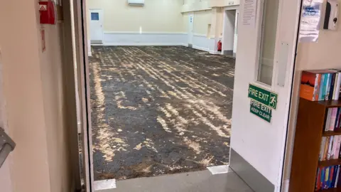 The picture shows the hall at Walsworth Community Center through the open fire doors. The flooring is completely damaged, with patches of black grit where the parquet once was. 