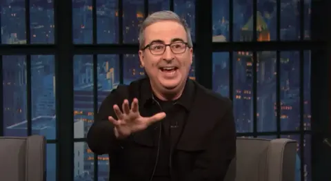John Oliver sits on a talk show chair speaking to camera - he is animated and reaches his hand out, gesticulating. He wears black glasses and has short grey hair and dark eyebrows.