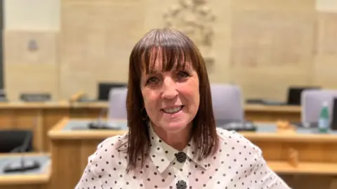 Lyn Worrall has shoulder length dark brown hair and a fringe smiling directly at the camera. She is wearing a white shirt with black poka dots. She is standing in a council chamber which is out of focus behind her.