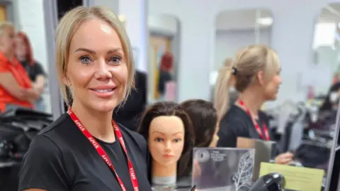 A blonde woman wearing a lanyard smiles at the camera. She is standing on front of a mirror which has a training mannequin head in front of it.