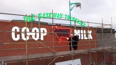 Central Co-op A sign with green lettering and lights saying "The Gateway to Health" and white lettering and lights saying "Co-op Milk". In the middle is a cow figure, which is lit up with red neon. It is on the side of a brick building 