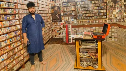 Abhishek Dey Vishal Kalita, wearing a blue kurta and holding his mobile phone in hand, poses near a shelf full of music cassettes in his library that has a collection of around 38,000 Zubeen Garg songs