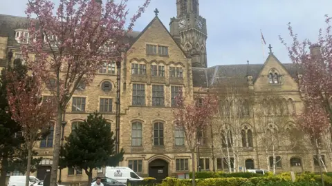 A grand historic stone building with a tall clock tower and pointed Gothic windows stands behind blooming pink cherry blossoms and green shrubs.