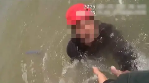 A blurred out face in water, he is wearing a black wetsuit and a red cap. There is a hand reaching out to get him out of the water.