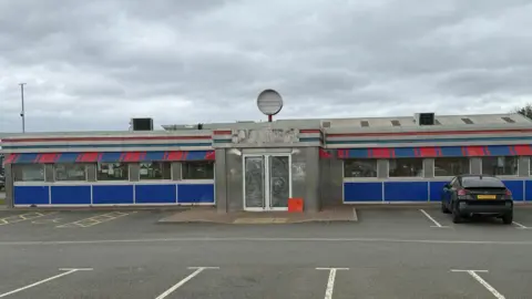 The former OK Diner building has the windows scrubbed and is in the process of shutting. It is a long single-storey concrete building with large windows and red and blue branding, in the style of a traditional 1950s American diner. A single black car is parked outside, to the right of the central entrance.