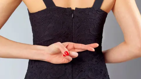 Getty Images A close of a woman wearing a black evening dress photographed from behind showing her waist and arms only . She has her fingers crossed behind her back and has red painted nails