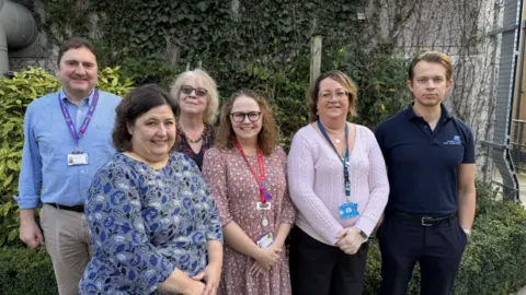 Somerset NHS A group of six people, men and women, all standing smiling to camera and wearing landyards. There is shrubbery behind them and what looks like a wire fence to the right.