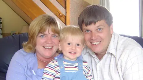 Family Photo James Evans at age four, he is sat in the middle next to Ruth and Andrew Evans. James is wearing a red, purple, blue, yellow stripey long-sleeve top. He is wearing blue dungarees and blonde hair. Ruth is wearing a blue top and cardigan. She has blonde hair which is curled at the ends. She is wearing a necklace. Ruth is smiling and she has blue eyes. Andrew is wearing a white shirt with grey squares. He is smiling and has brown hair and blue eyes. 