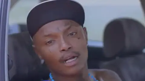 3Dimensions SA Shebeshxt sitting in a car with blue necklace and a cap on