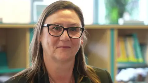Manda George has long, light brown hair and is wearing a dark green suit jacket and glasses