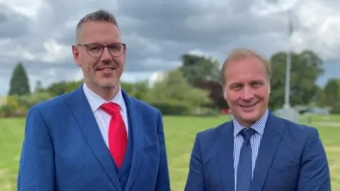 Two men in blue suits, one with a red tie and the other with a blue tie standing in a grassy field