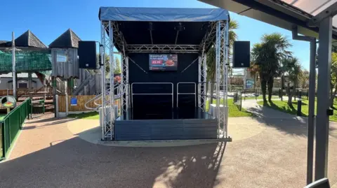 Parkdean Resorts A small outdoor stage can be seen, with metal supports and a covered roof in the foreground in front of a children's play park