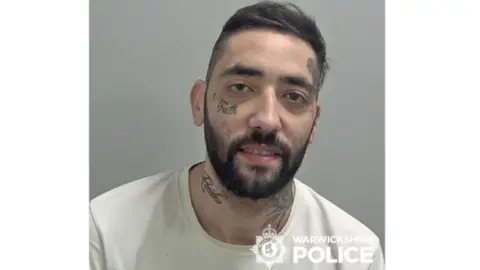 A mugshot of James White looking at the camera. He has a dark-haired beard with a tatoo under his eye, on the right side of his face above his eyebrow, and two tattoos on his neck.