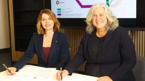 Kim McGuinness and Heidi Alexander sit side by side at a desk while signing the document. They are both looking at the camera and smiling. McGuinness, on the left, has mid-length blonde hair and is wearing a blue blazer. Alexander has mid-length light blonde/grey hair and is wearing a blue blazer.