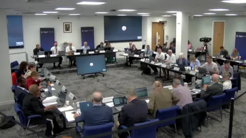 BBC A picture of Cherwell District Council's planning committee. Councillors and officers sit in a square of desks around a central screen, with other screens on the walls. Members of the committee are also using laptops.
