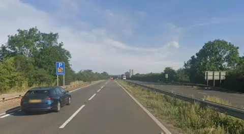 The A1. The road, which stretches into the distance, is almost empty. Each carriageway has two lanes. There is a blue estate car in the foreground on the left of the picture, heading away from the camera. In the far distance, heading towards the camera, there is white lorry. It is a clear day and there are leaves on the trees beside the road.