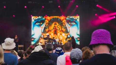 Holly Bradley Cast performing at Bristol Sounds. The picture is taken from the crowd. Backs of people's heads can be seen and the band is in the distance. The stage is orange with two blue faces projected on the back.