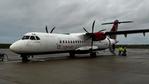 A Loganair plane on the tarmac at Jersey Airport on a cloudy day. The aircraft is white and red. It has black twin propellers. Two people in high-vis jackets are at the back of the plane.