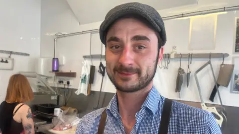 Butcher Nicholas David stands in his shop. There are knives and instruments behind him.