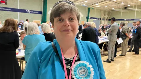 A woman with short brown hair, a blue jacket and a blue Reform UK rosette stands and smiles in the middle of a sports hall as an election count takes place behind her.