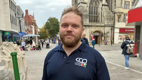 Richard Stone stands in the centre of Gloucester. He is wearing a blue polo top with a colourful logo just below his left shoulder