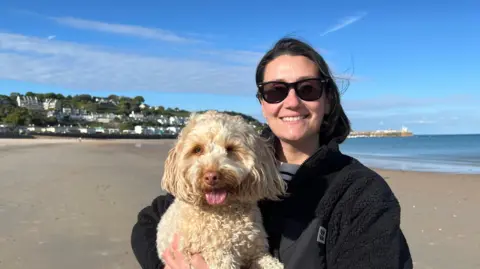 Annabel Hill wears dark sunglasses and a black fleece jacket and stands on a sandy beach holding a small, curly‑haired dog. The dog has light-coloured fur and its tongue is visible. Behind them is a calm sea, a clear blue sky with scattered clouds, and white buildings on a hillside. A pier extends into the water in the distance.